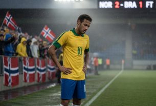 Neymar stands alone on a rain-damp football pitch under stadium lights, head bowed in